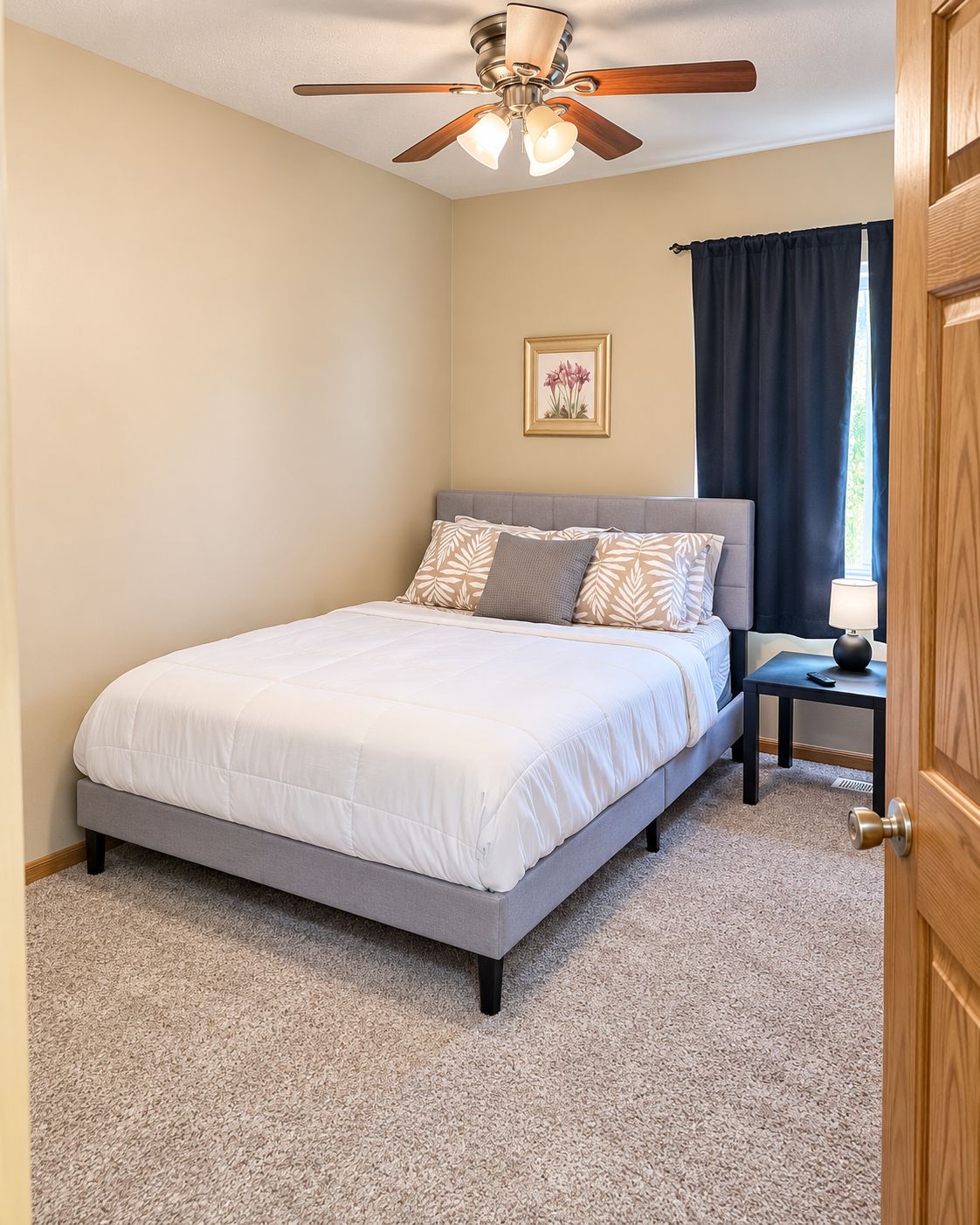 Une chambre à coucher comprenant un lit, une table de chevet, un ventilateur de plafond, un tableau encadré accroché au mur et une fenêtre avec des rideaux.