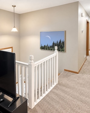 A hallway with a white railing, a mounted picture of a crescent moon and trees, and a television on a stand. - Serene 3-bedroom 2-bath house (Omaha)