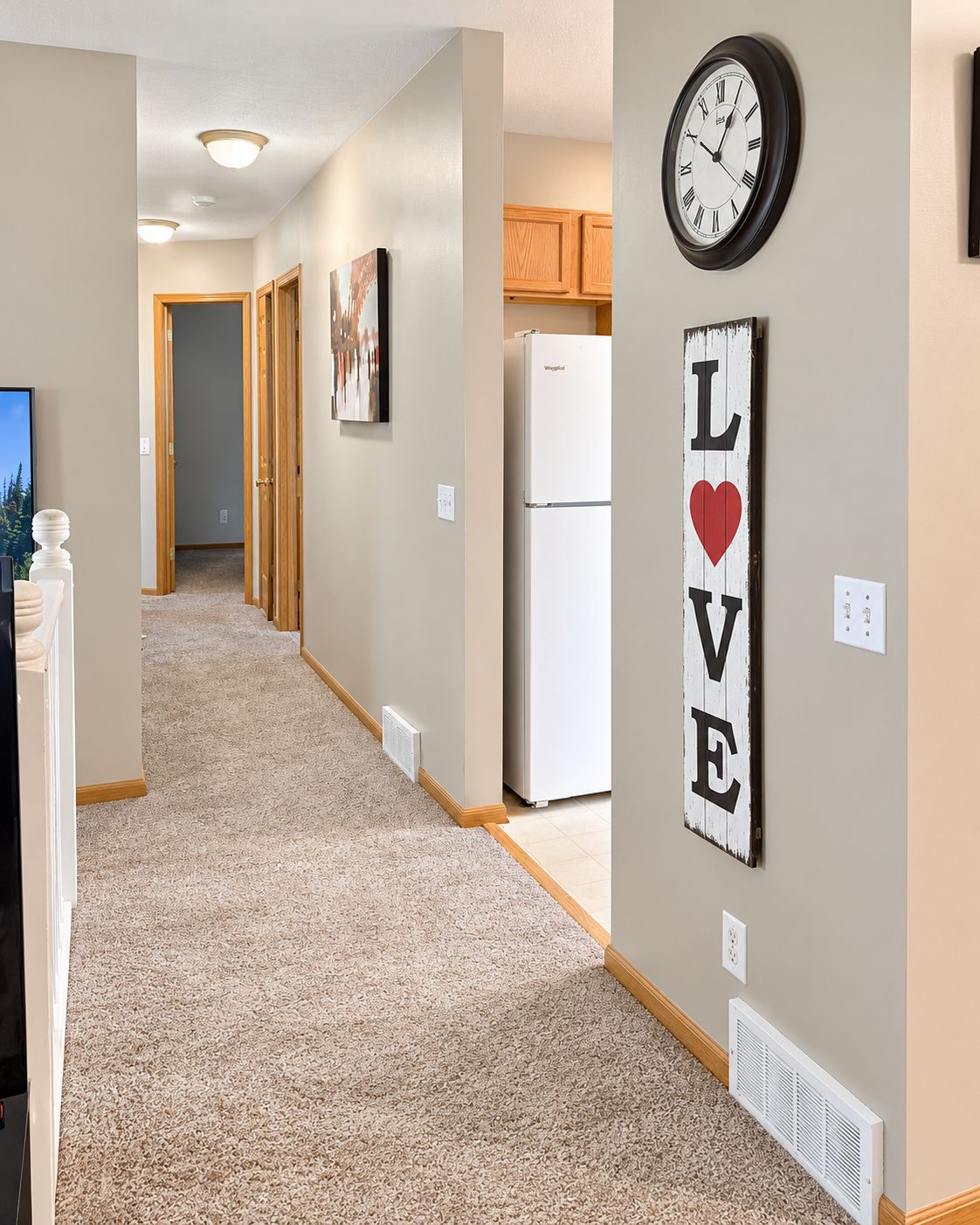 A hallway with carpeted flooring, a wall-mounted clock, a refrigerator, and a 'LOVE' sign.