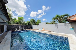A swimming pool with a tiled edge, a stone wall, and a palm tree in the background. - Ava Guest House Bali (Ungasan)