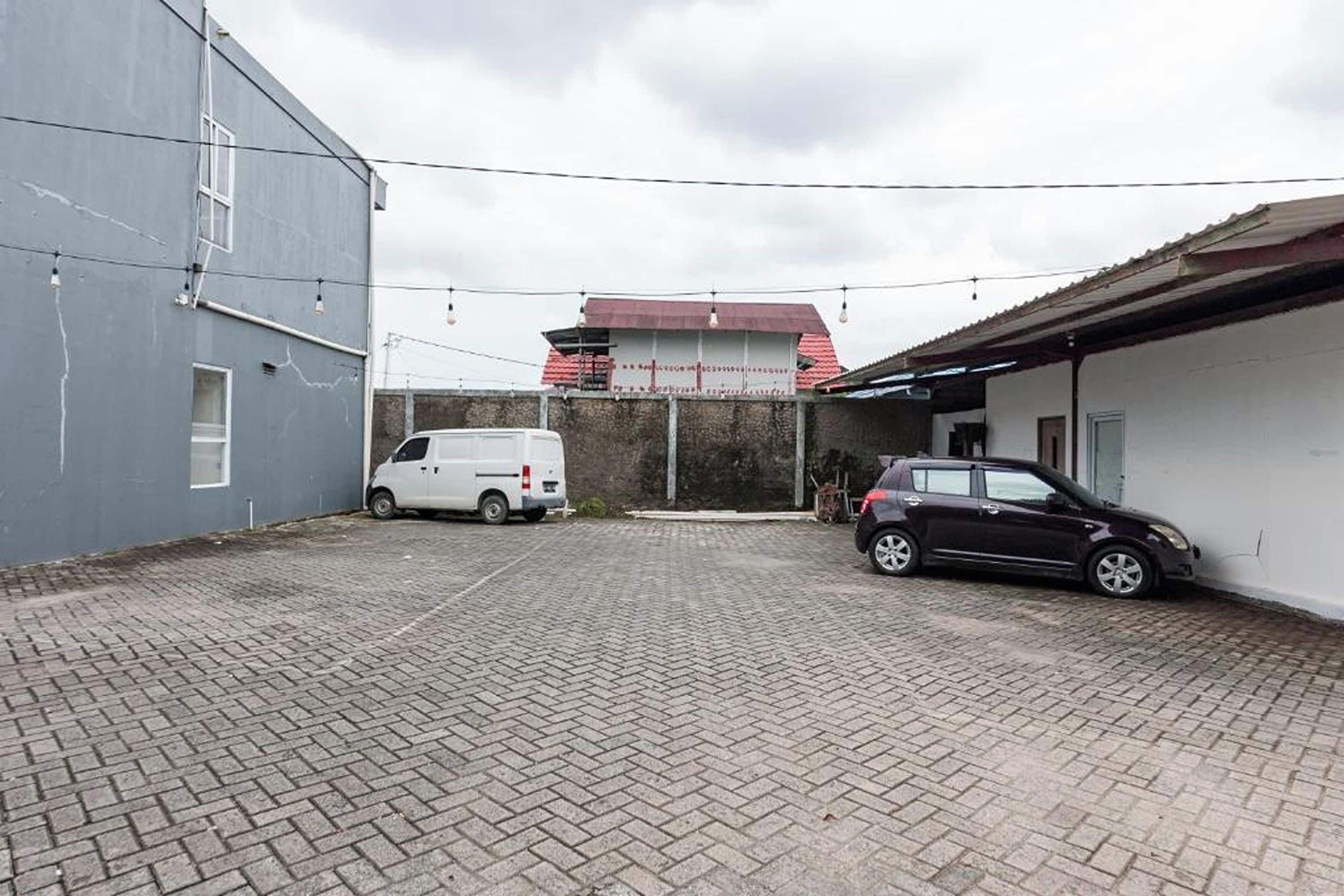 A paved courtyard with a white van and a dark car parked, a building with a red roof, and a grey wall with a window.