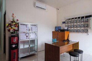 A hotel room with a wooden desk, a glass cabinet with towels, a red vending machine, a flower arrangement, and a wall-mounted electrical panel. - Ava Guest House Magelang (Magelang)