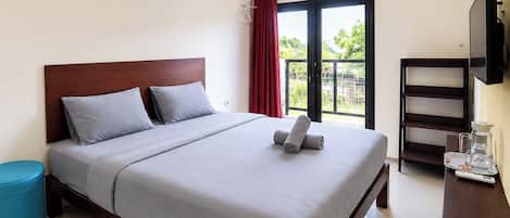 A hotel room with a bed, a small blue ottoman, a TV, and a window with a view of greenery.