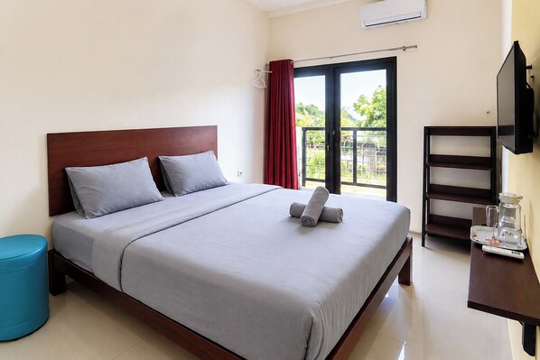 Habitación de hotel con cama, un pequeño taburete azul, televisor y ventana con vistas a zonas verdes.