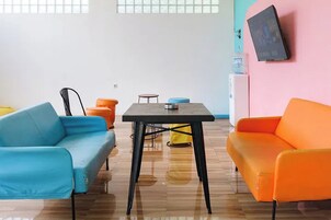 Una habitación con piso de madera, un sofá azul, un sofá naranja, una mesita redonda y un televisor montado en la pared.
