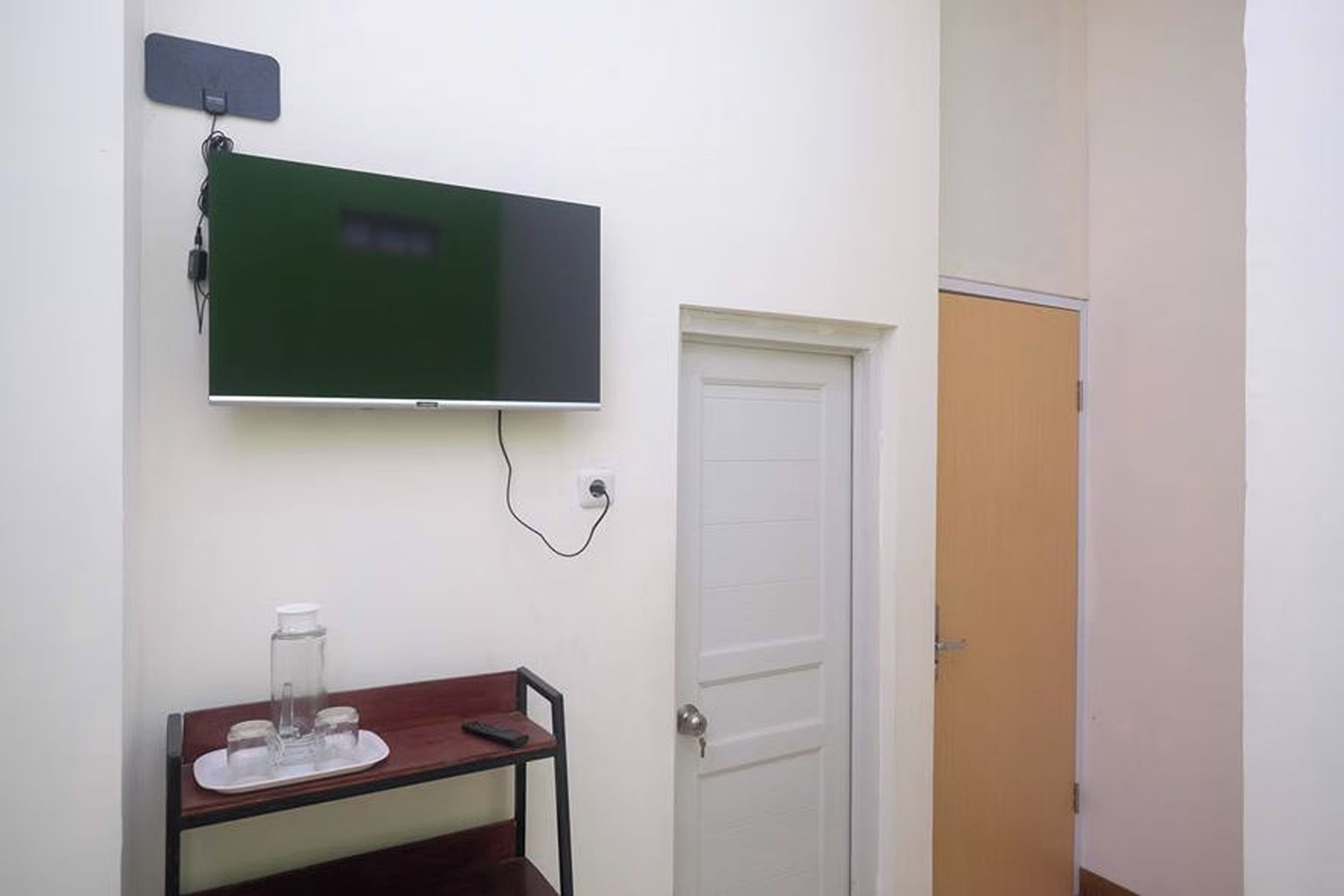 Quarto com televisão na parede, porta branca e pequena mesa com um recipiente de vidro.