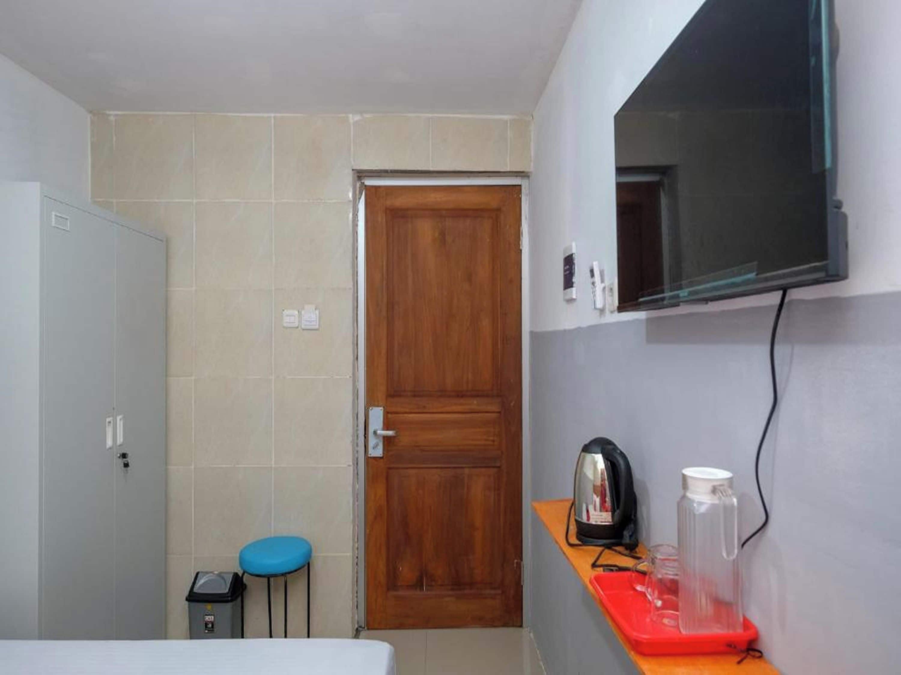 A small room with a wooden door, a television mounted on the wall, a small table with a kettle and a glass container, and a blue stool.