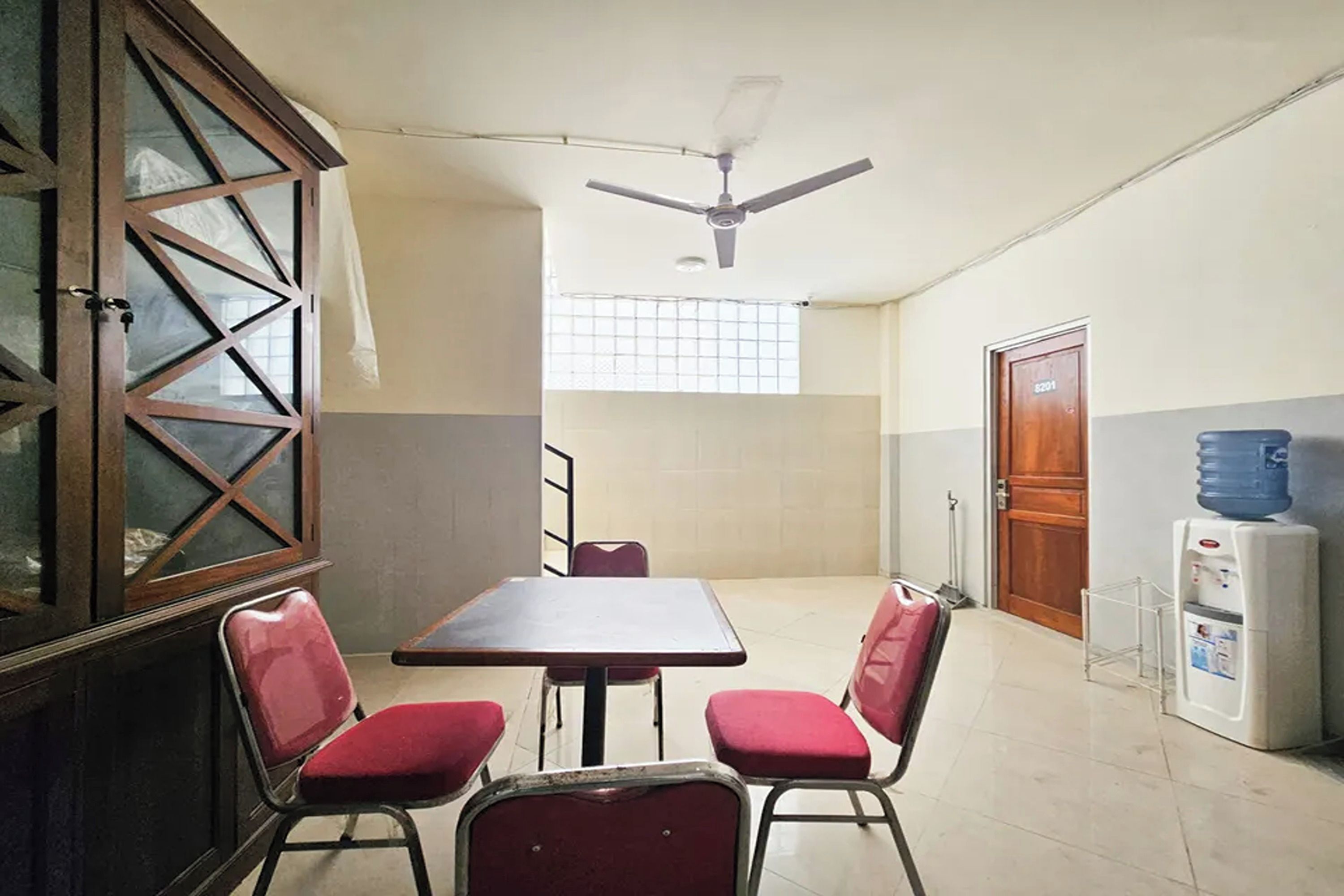 Una habitación con una mesa, cuatro sillas, un ventilador de techo, una ventana con persianas y un dispensador de agua.