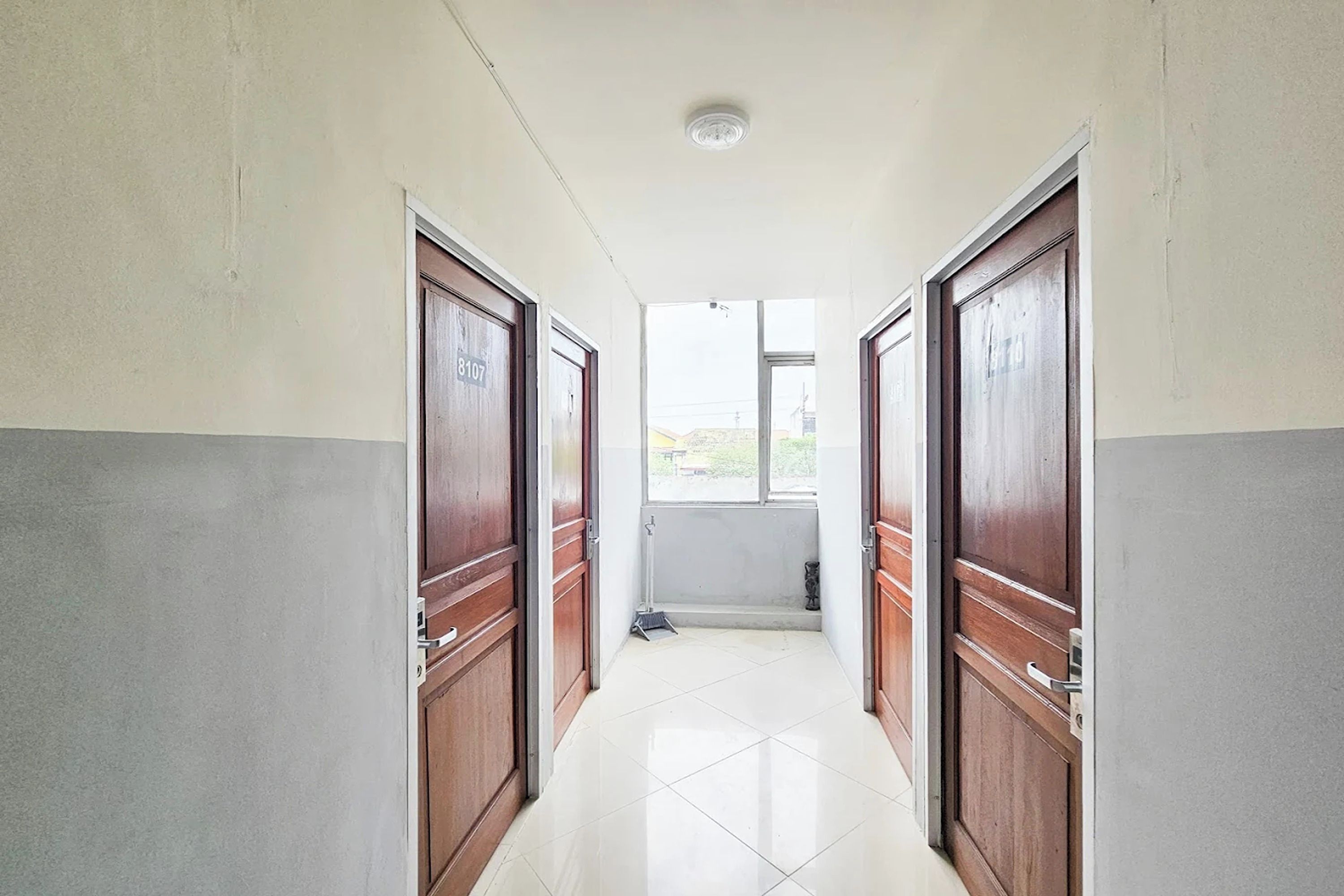 Un couloir avec deux portes en bois, un sol carrelé de carreaux blancs et une fenêtre au fond.