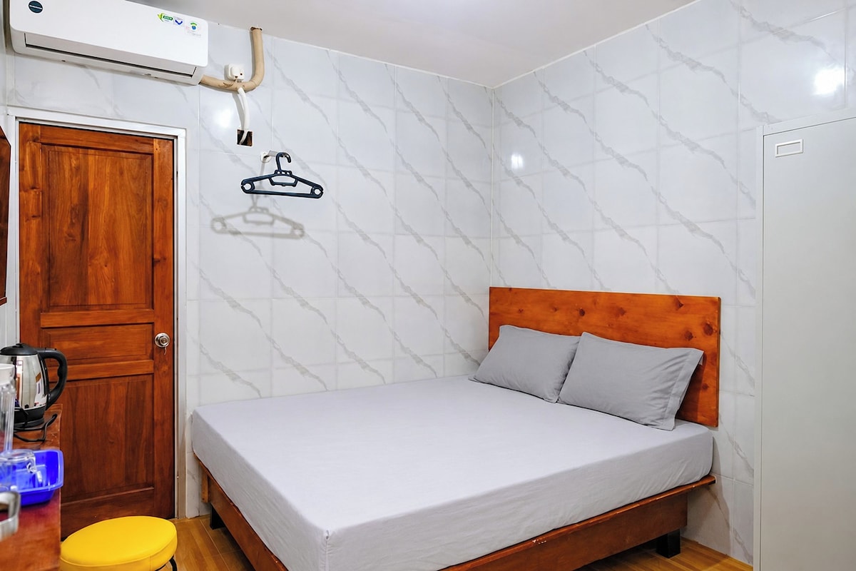 Een slaapkamer met een houten bedframe, een witte matras en een houten hoofdbord. Er is een houten deur en een klein, geel krukje.