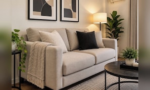 A beige sofa with a throw blanket and two pillows, a floor lamp, and a small round table with candles. - The Orabella Residences - Qc (Cubao) (Quezon)