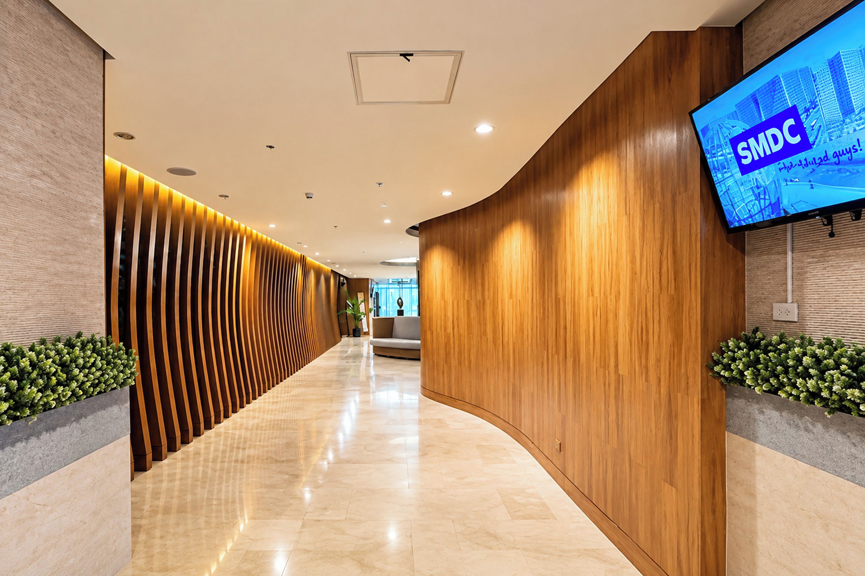 Un hall d’entrée moderne avec un mur en bois aux lignes courbes, un téléviseur à écran plat diffusant le logo « SMDC », et un sol en marbre.