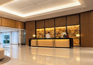 A hotel reception area with a marble wall, a reception desk, and a clean, reflective floor. - Shell Residences Cozy Staycation (Pasay)