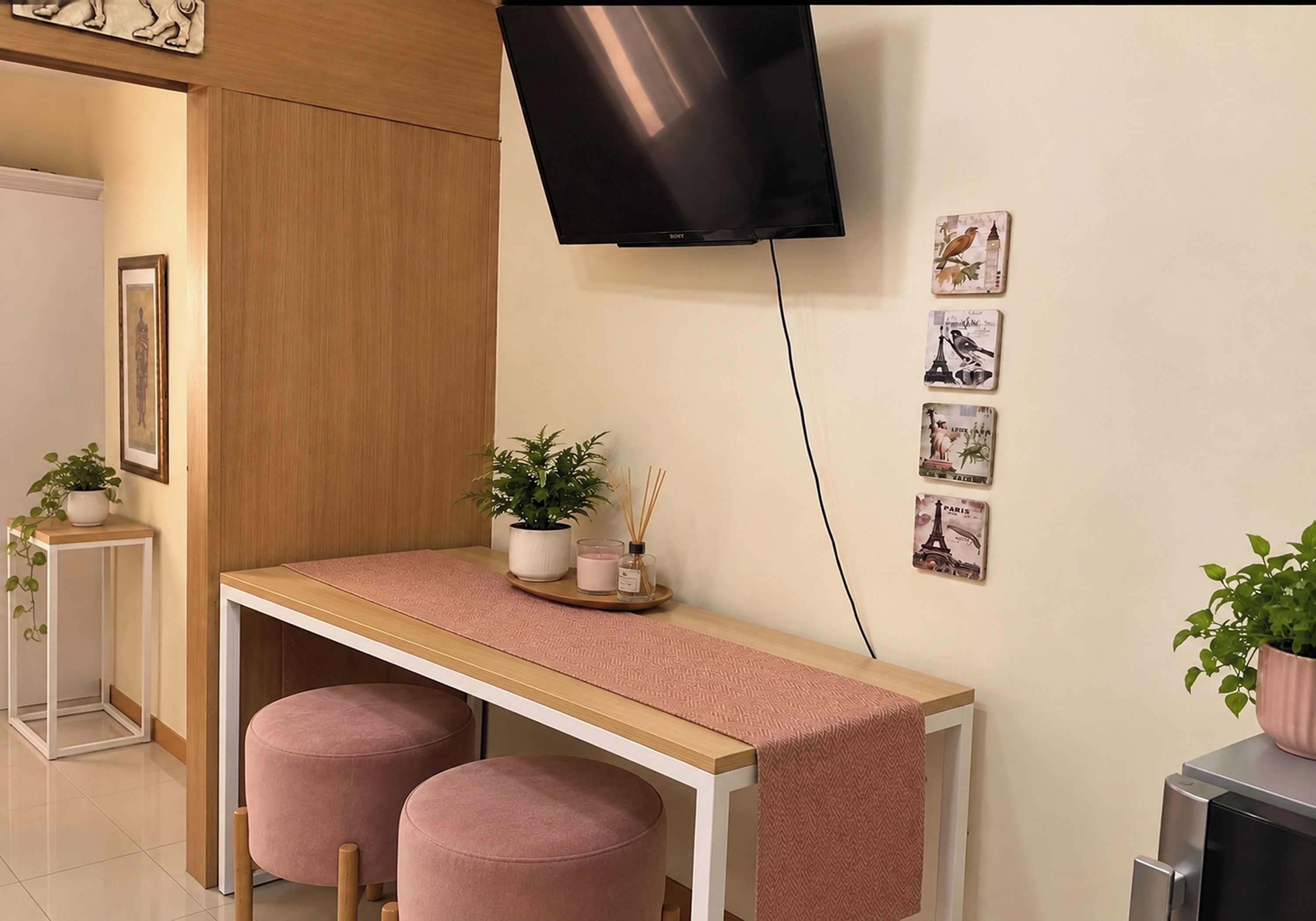 A modern room with a flat-screen TV mounted on the wall, a small table with a pink cushion, and a potted plant.