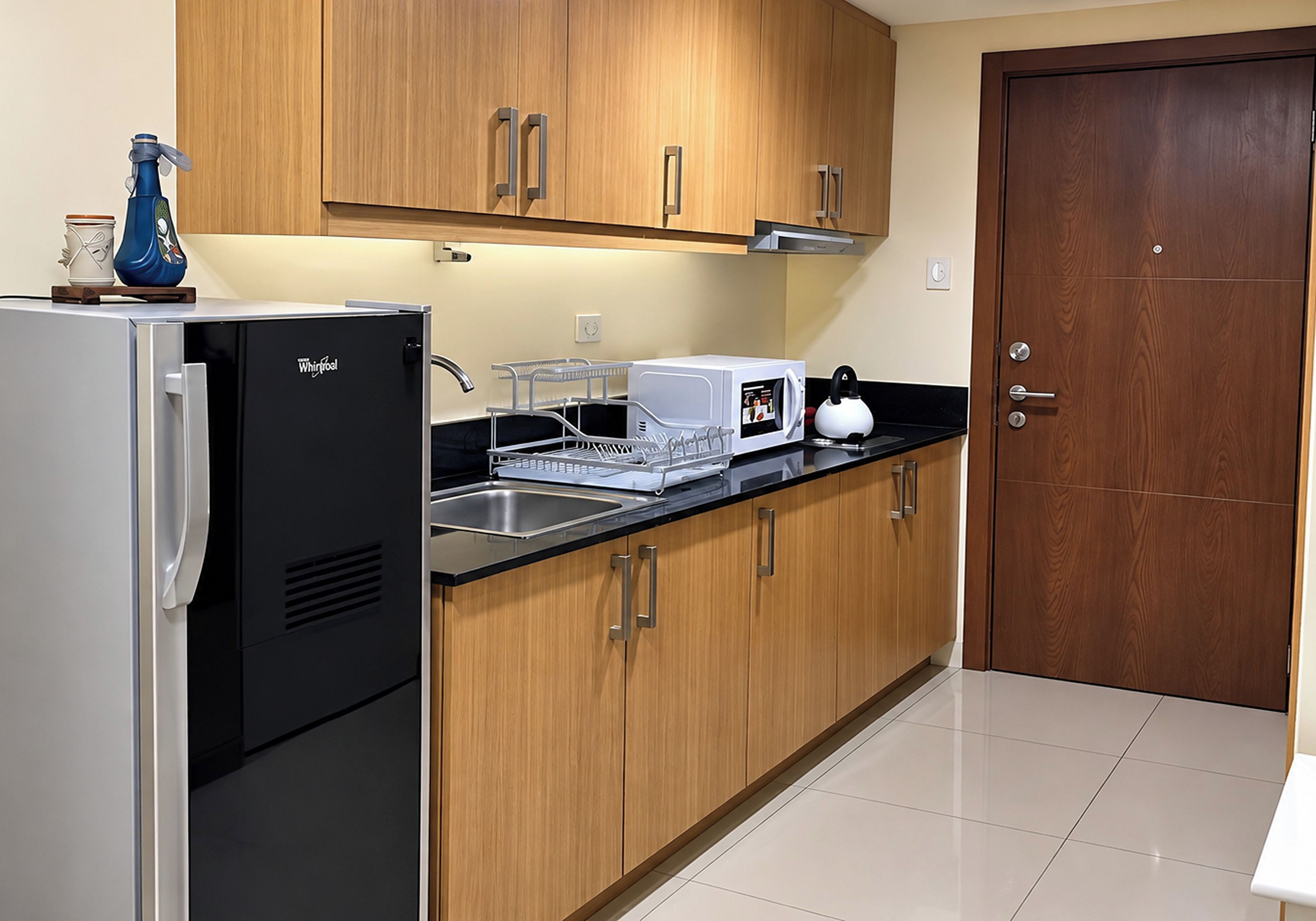 A kitchen with a refrigerator, microwave, kettle, and wooden cabinets.