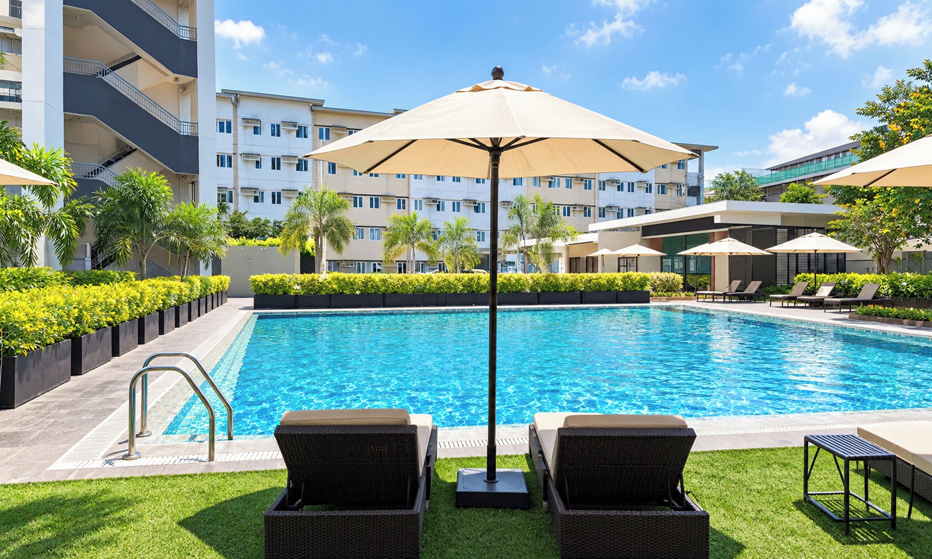 Ein Swimmingpool mit Liegestühlen und Sonnenschirmen, umgeben von Grünflächen und einem mehrstöckigen Gebäude.