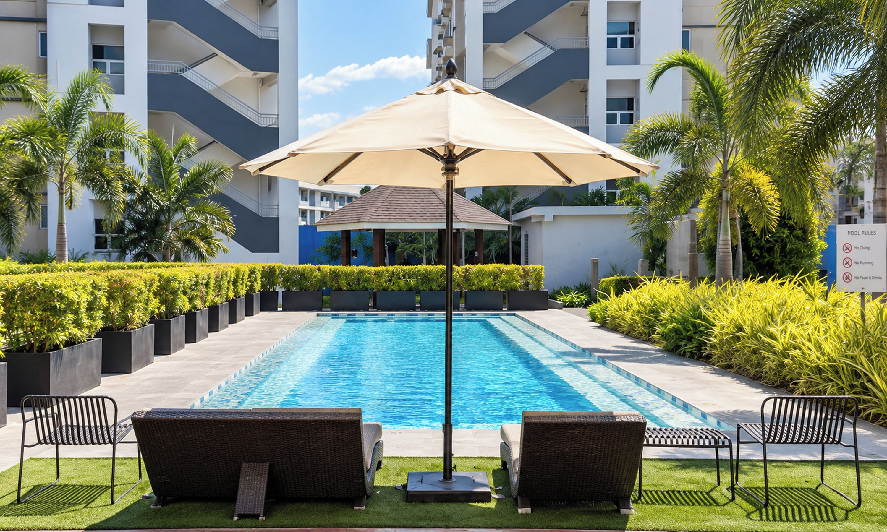 A swimming pool with lounge chairs and an umbrella, surrounded by greenery and a multi-story building.