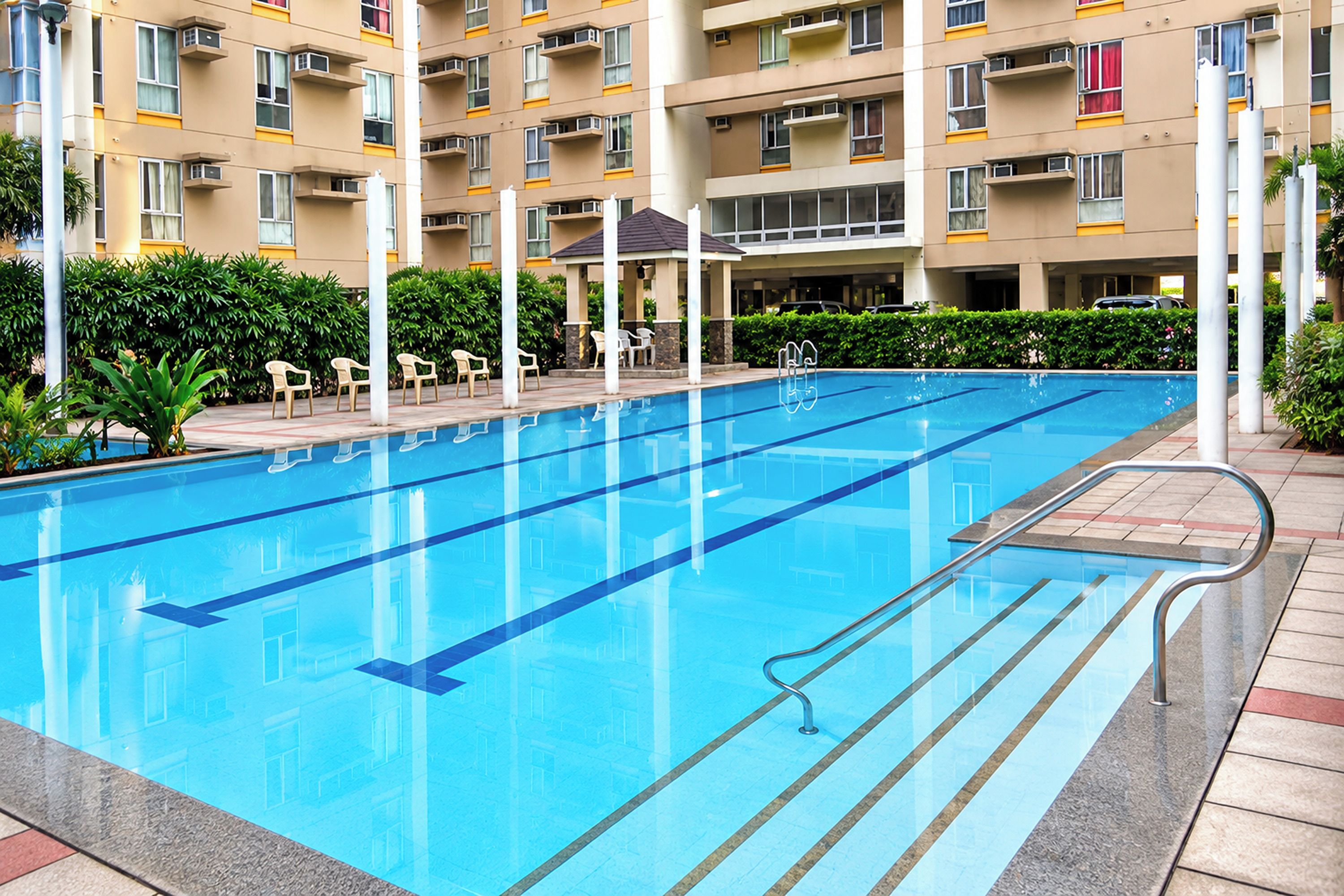 A swimming pool with clear blue water, surrounded by a tiled deck and a building with multiple balconies in the background.