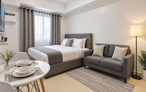 A hotel room with a bed, a sofa, a dining table set for two, and a lamp. - Tagaytay Serin West Studio (Tagaytay)