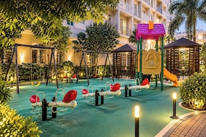 A children's play area with swings and a slide, surrounded by trees and a pool. - Naia Smdc South Residences (Las Pinas)