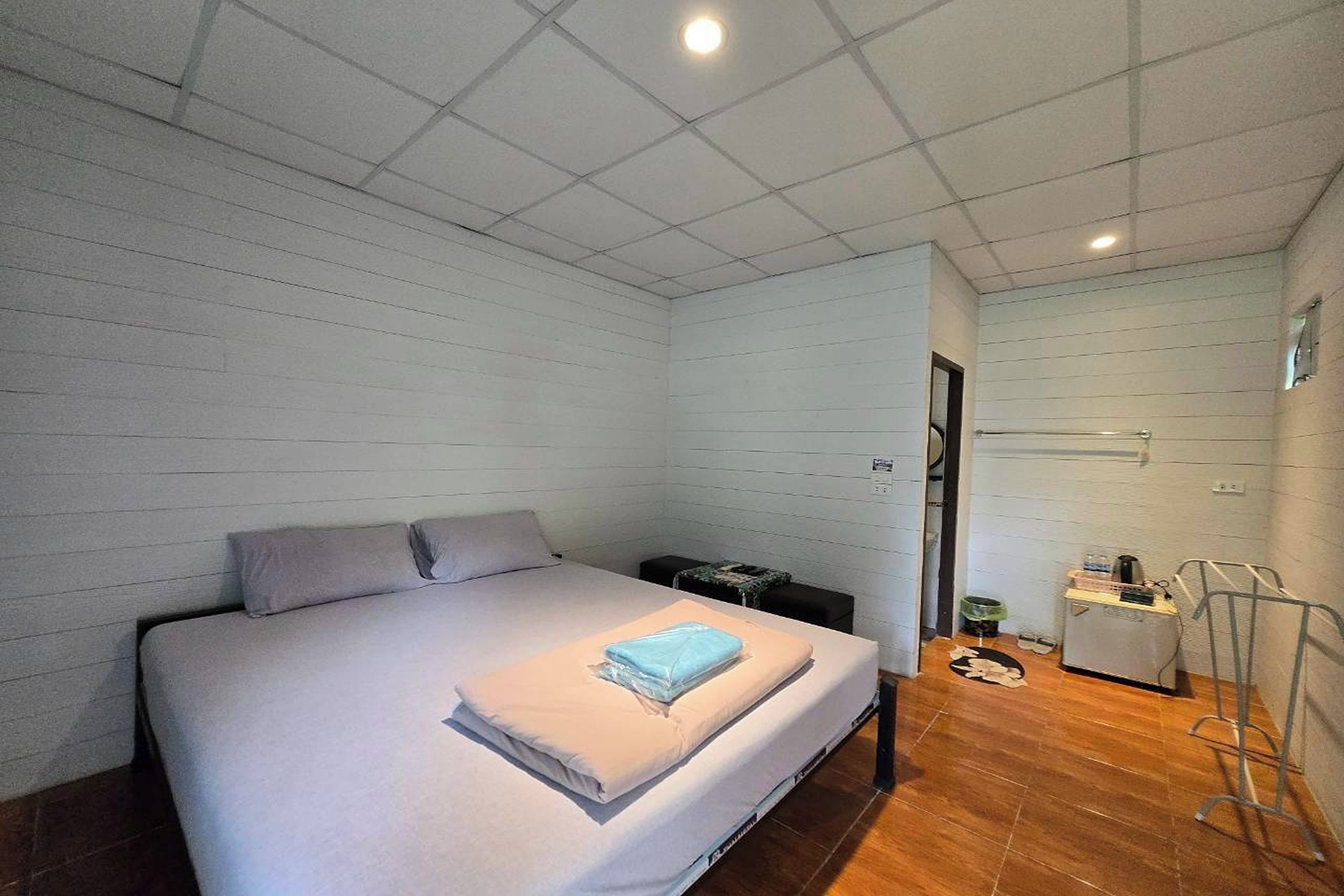 A single-bed room with a wooden floor, a bed with a white mattress and pillows, a small bedside table, and a wall-mounted shelf.
