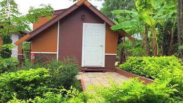 A small, single-story building with a brown exterior and a white door, surrounded by lush greenery.