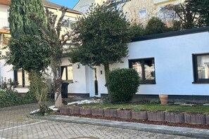 Un immeuble résidentiel à la façade blanche, doté d’un petit jardin avec un arbre et un arbuste, et d’une allée pavée.
