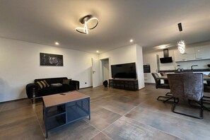 A modern living room with a sofa, coffee table, and TV. There is a dining area with chairs and a table. The kitchen is visible in the background. - Holiday home with 2 bedrooms (Lahnstein)