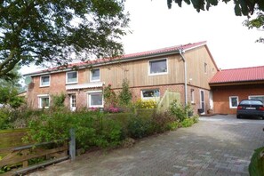 Une maison à deux étages, avec une façade en briques, un toit recouvert de tuiles rouges et une allée pavée menant à une voiture garée.