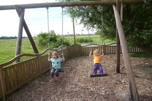 Two children on swings in an outdoor play area with a wooden fence and trees in the background. - Holiday apartment with 2 bedrooms (Poppenbüll)