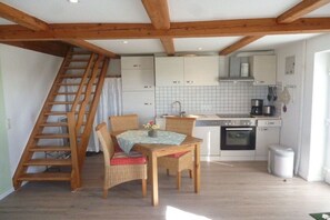 A kitchen with white cabinets, a wooden staircase, a dining table with chairs, and a small trash bin. - Holiday apartment with 2 bedrooms (Poppenbüll)