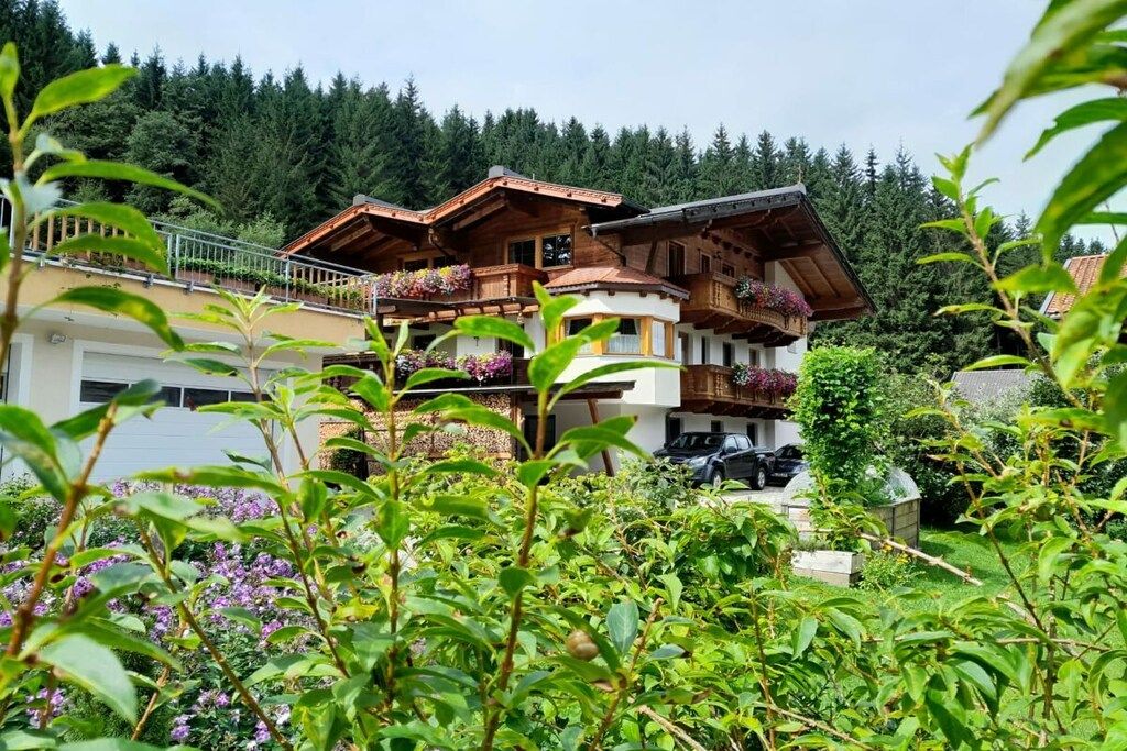 Een meerlaags chalet met balkons, omgeven door weelderige begroeiing en een bebost achtergrond.