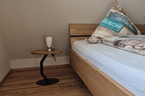A bedroom with a wooden bed, a small round table with a lamp, and a pillow with a beach-themed design. - Holiday apartment with 1 bedroom (Norden-Norddeich)