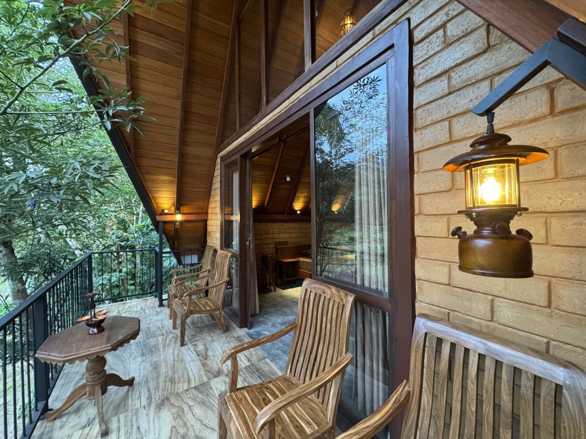 A wooden balcony with chairs and a table, a lantern, and a view of trees.