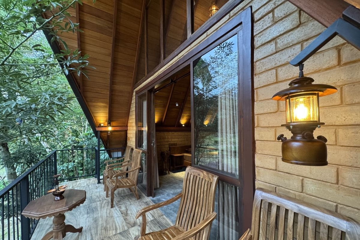A wooden balcony with chairs and a table, a lantern, and a view of trees.