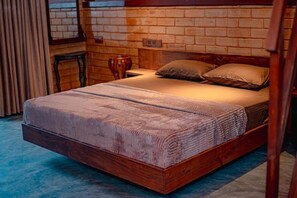 Un lit simple avec un cadre en bois et une couvre-lit à motifs, placé contre un mur de briques avec une fenêtre et des rideaux.