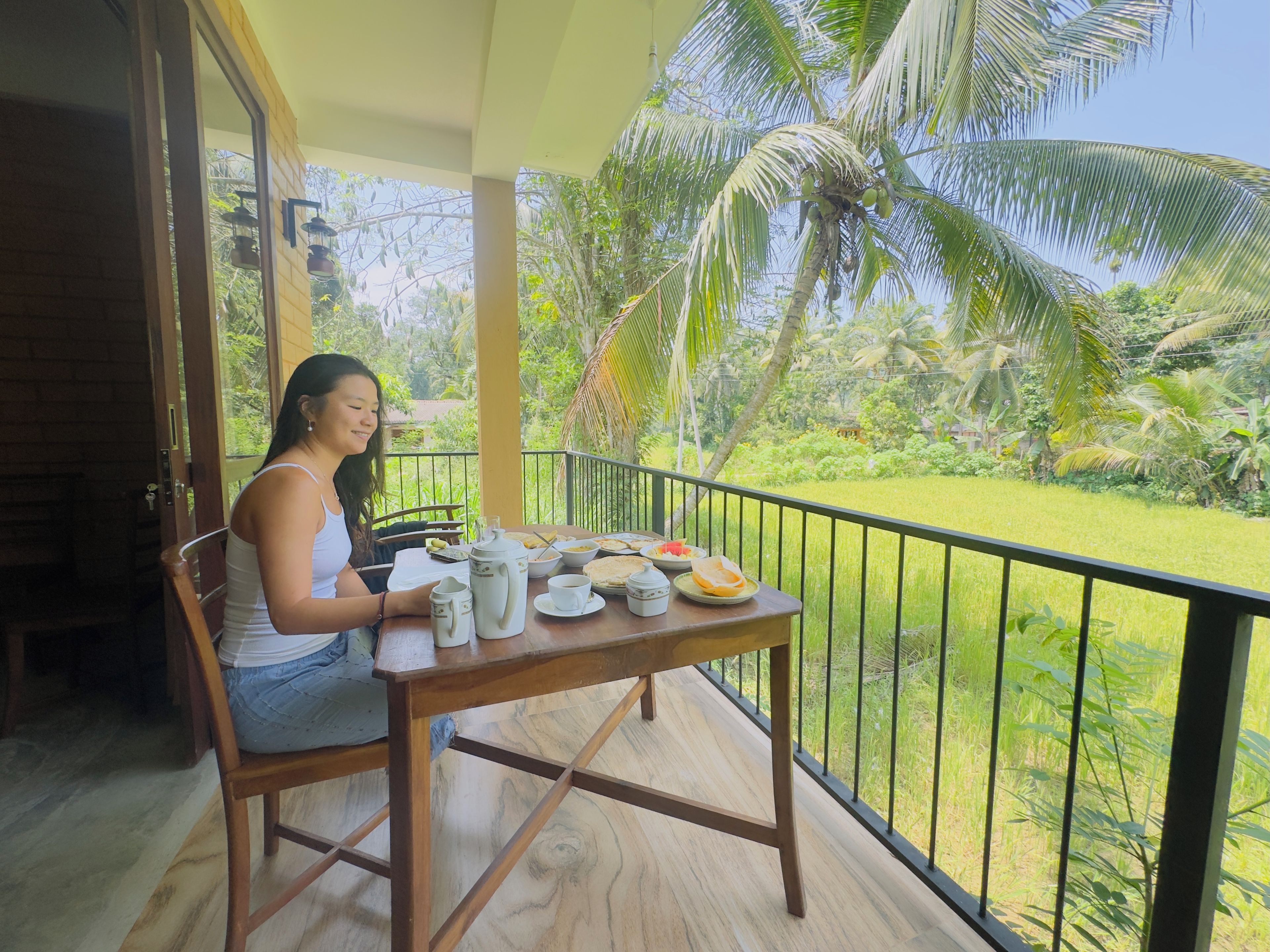 Una persona sentada en una mesa de madera en un balcón con vistas a un exuberante jardín y palmeras.
