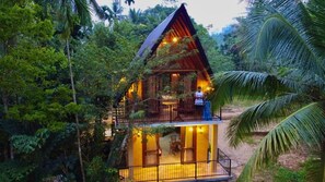 Una baita di legno a due piani con un balcone, immersa in una fitta vegetazione tropicale.