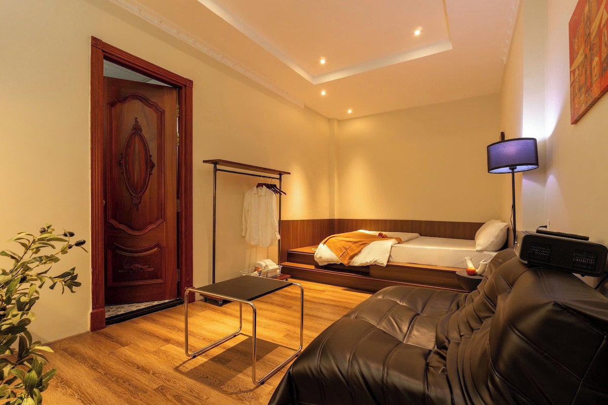 Une chambre d’hôtel avec une porte en bois, un lit avec une tête de lit, un canapé en cuir noir, une petite table et une lampe fixée au mur.