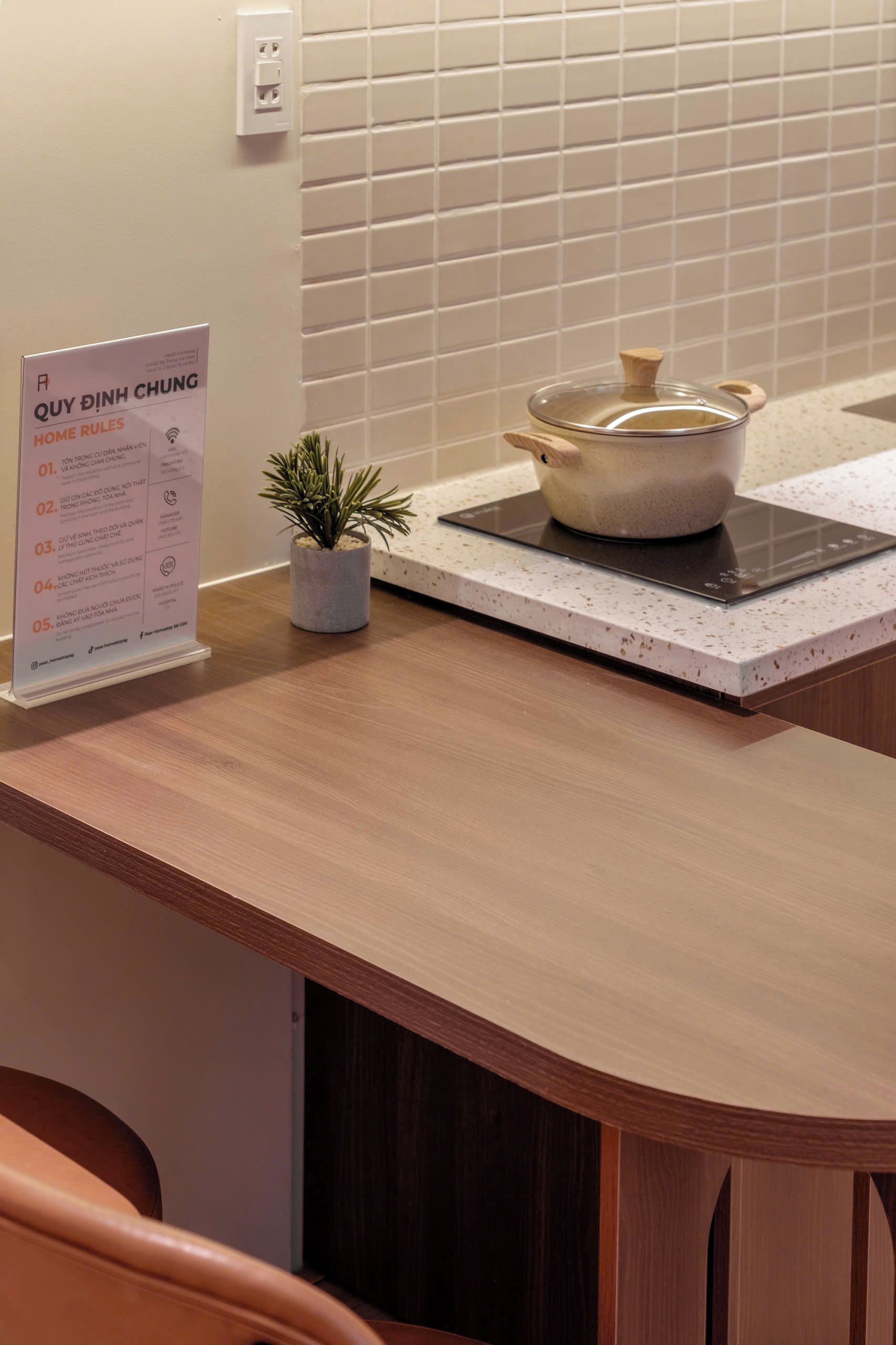 Una encimera de cocina con una olla sobre una cocina de inducción, una pequeña planta en maceta y un cartel con normas de la casa.