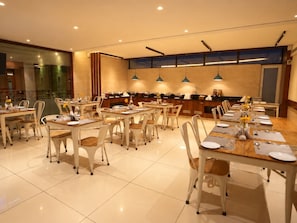 Comedor de hotel con varias mesas y sillas, una cocina al fondo e iluminación colgante.