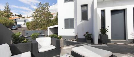 A modern outdoor seating area with wicker furniture and a glass table, surrounded by potted plants and a white building with a grey door.