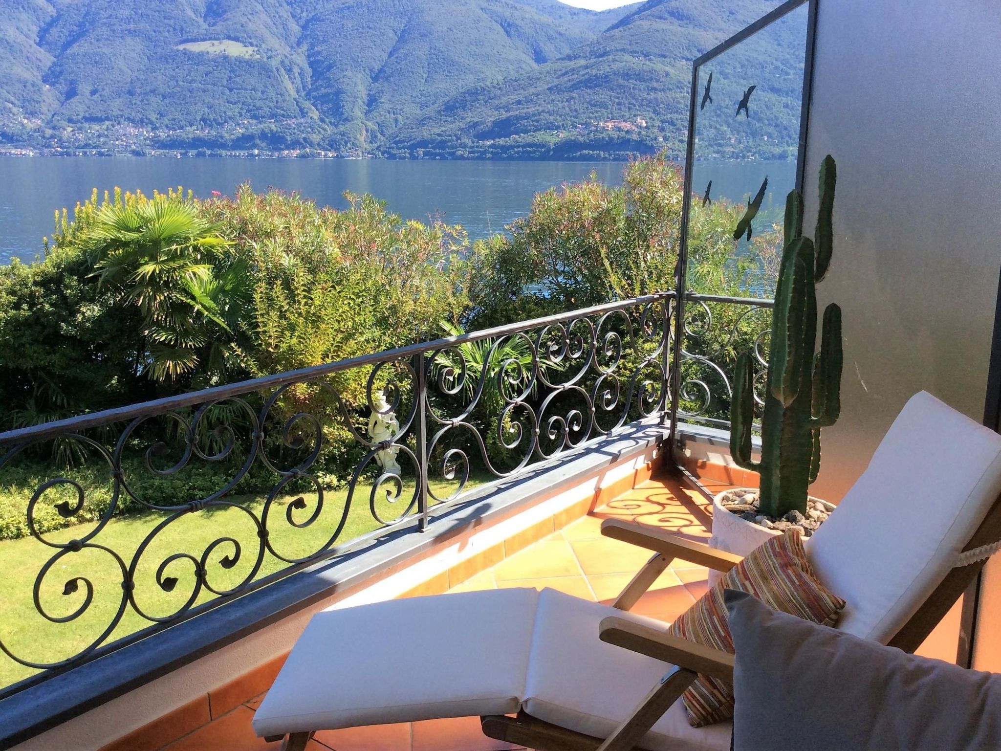 Een balkon met uitzicht op een meer en bergen, twee lounge stoelen en een cactus.