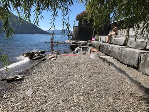 A pebbled beach with a rocky shoreline, a stone wall, and a person sitting on the wall. - La Palma by Interhome (Brissago)