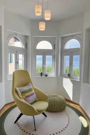Ein Zimmer mit einem gelben Sessel, einem runden gewebten Pouf und drei Pendelleuchten. Es gibt drei Bogenfenster mit Blick auf das Meer.