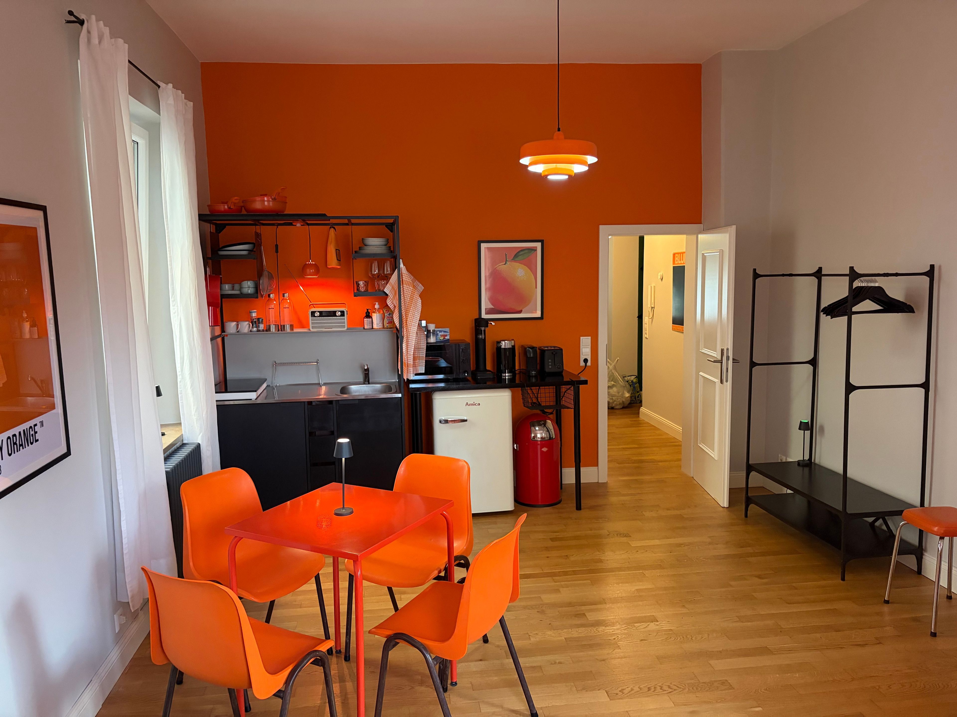 Ein kompakter Wohnbereich mit Küchenzeile, einem Essbereich mit orangefarbenen Stühlen, einer roten Gasflasche und einem schwarzen Regalelement.