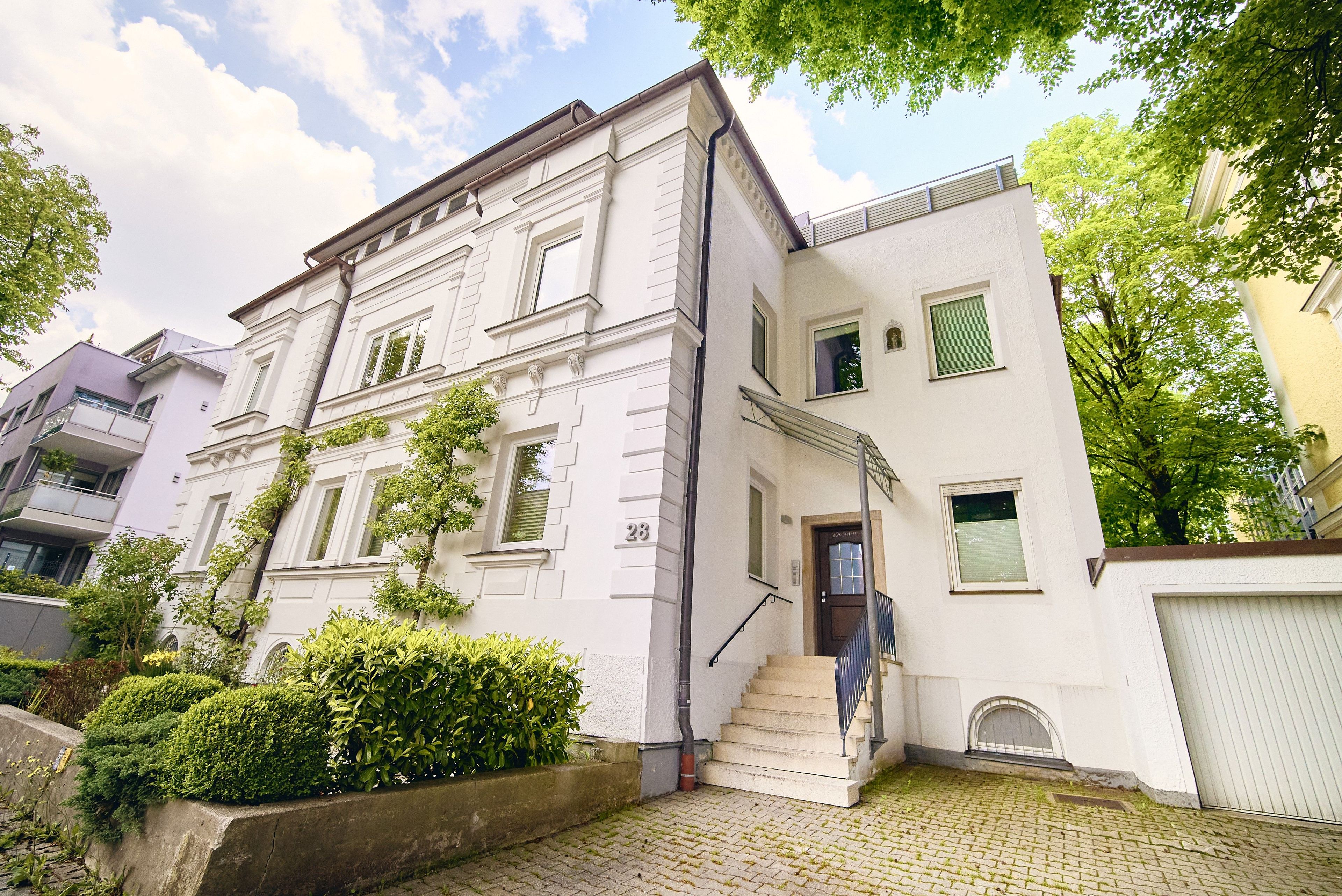 Ein mehrstöckiges Gebäude mit weißer Fassade, einer sichtbaren Treppe und einem kleinen Gartenbereich mit Hecken.