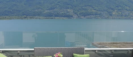Una terrazza con mobili in vimini e ringhiera in vetro che si affaccia su un lago e montagne.