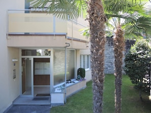 A modern two-story building with a glass door entrance, a small garden area with palm trees, and a stone wall in the background. - Attico Luciana by Interhome (Minusio)