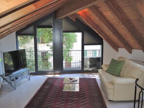 A spacious attic room with a large TV on a stand, a white sofa, a patterned rug, and a view of greenery through large windows. - Casa Gialla by Interhome (Locarno Tegna)